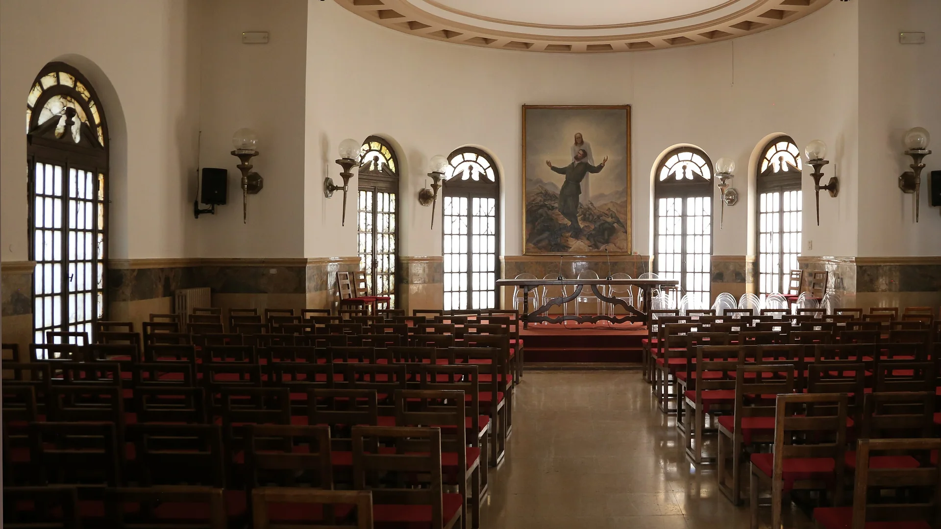The Sala Antonio Girelli in the Palazzo dei Mutilati, Verona: rows of wooden chairs with red seats facing a curved apse with arched windows and a large painting.